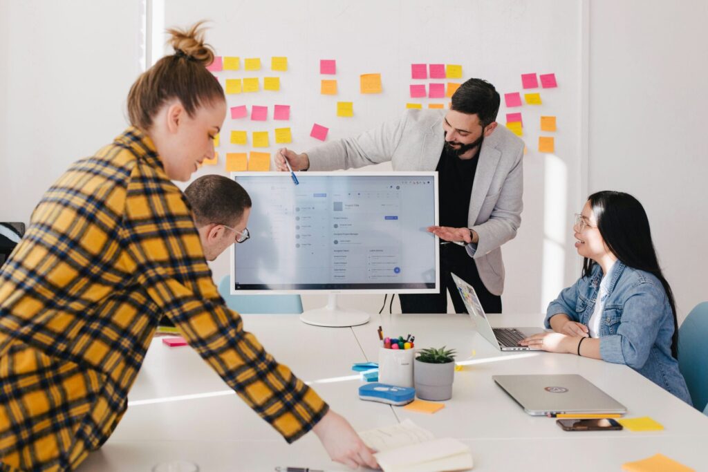 Four people working together at a table. One man is pointing out information on a screen. There are colourful sticky notes on the wall.